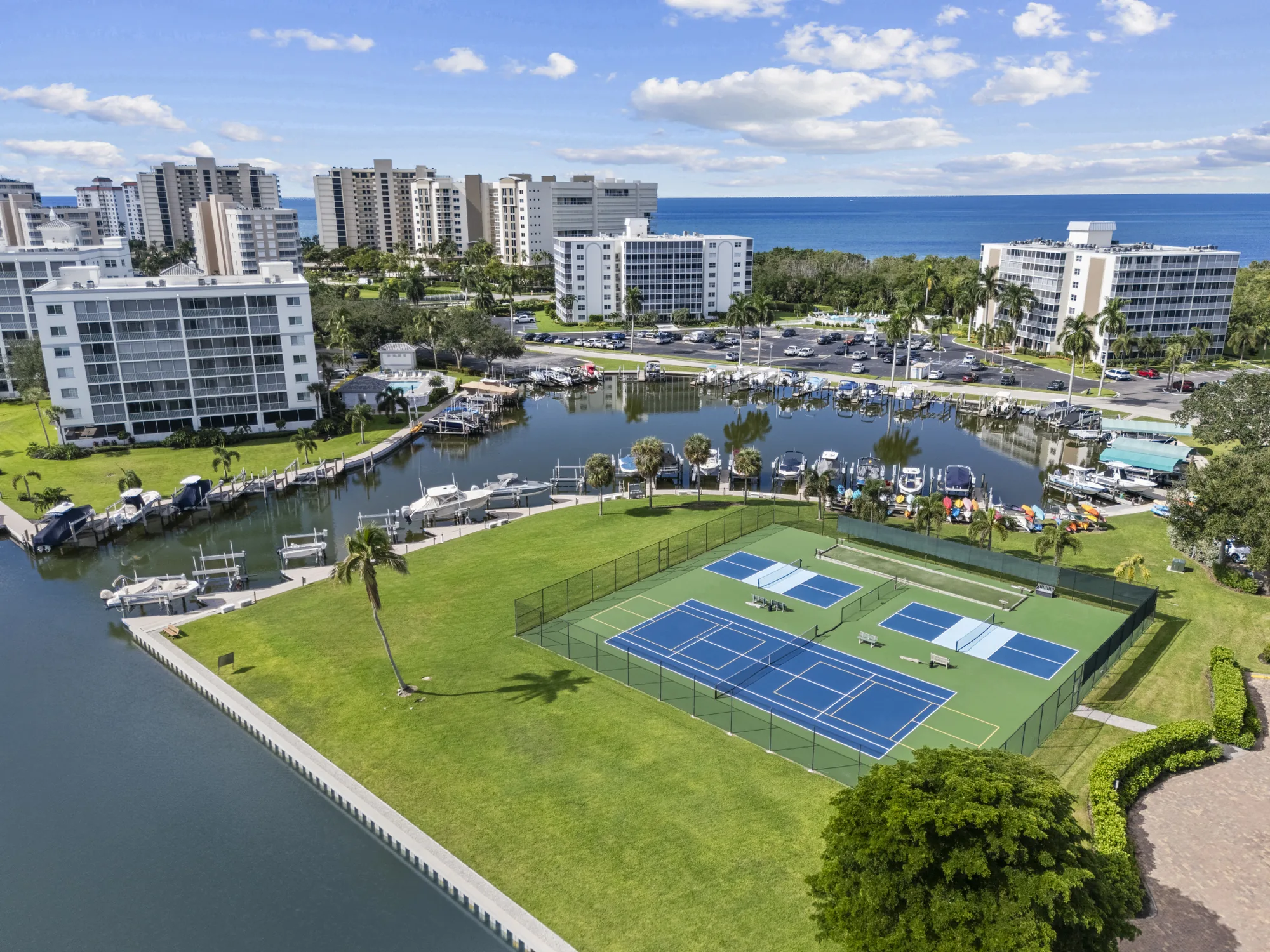 Aerial of the marina basin on the Bluebill peninsula at Vanderbilt Beach, with boat docks and lifts, tennis and pickleball courts and the Gulf of Mexico beyond, Naples, Florida