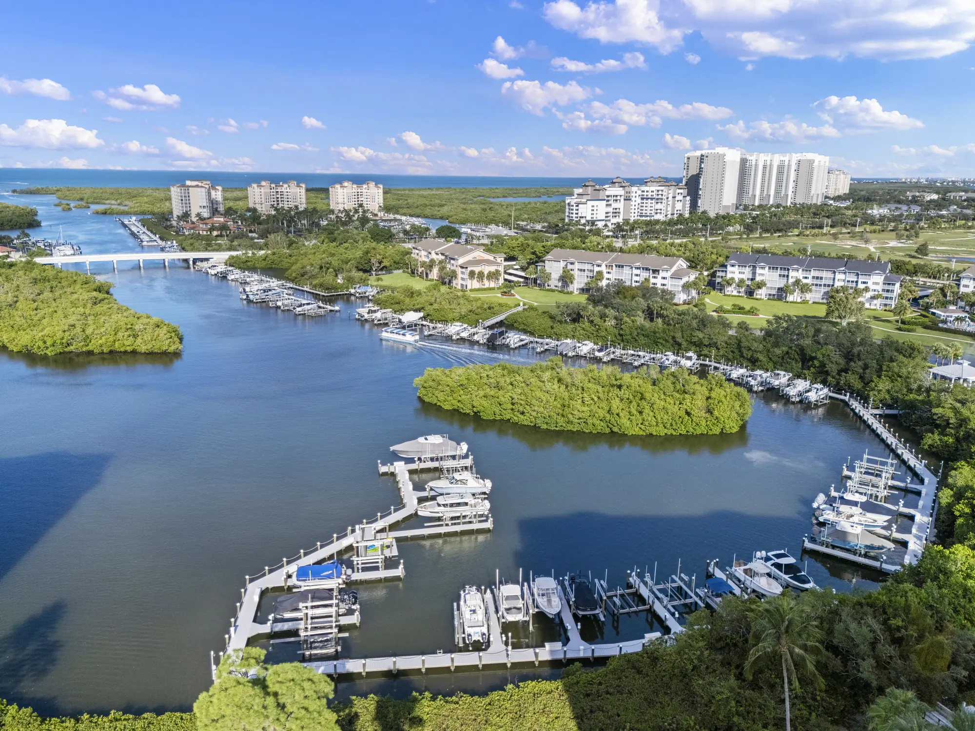 Aerial of the Wiggins Bay marina beside Cove Towers, with its docks and boat lifts on the bay and Wiggins Pass beyond, North Naples, Florida